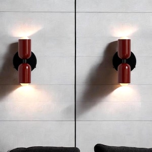 Könnte beinhalten: Zwei rote und schwarze Wandleuchten im modernen Design. Jede Leuchte hat zwei Glühbirnen und eine zylindrische Form.