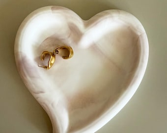 Könnte beinhalten: Eine herzförmige Schmuckschale mit einem marmorierten weiß-rosa Design. Ein Paar goldene Creolen liegt in der Schale. Die Schale besteht aus einem festen Material und hat eine glatte Oberfläche.