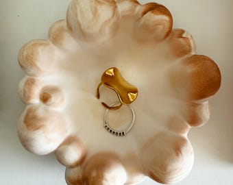 Könnte beinhalten: Dekorative, wolkenförmige Keramikschale in Creme- und Brauntönen. Die Schale enthält drei Ringe: einen goldfarbenen Ring, einen silbernen Ring mit kleinen schwarzen Akzenten und einen dünnen goldenen Ring. Einzigartiges, strukturiertes Design.