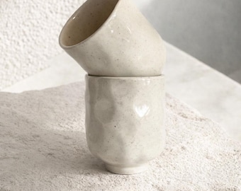 Könnte beinhalten: Zwei cremefarbene Keramikbecher, die auf einer strukturierten, hellen Oberfläche gestapelt sind. Die Becher haben ein gesprenkeltes Aussehen und ein leicht unregelmäßiges, facettiertes Design. Der obere Becher ist geneigt.