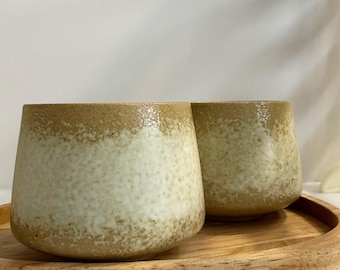 Könnte beinhalten: Zwei Keramikbecher mit einer gesprenkelten, cremefarbenen und beigefarbenen Glasur. Die Becher haben eine abgerundete Form und stehen auf einem runden Holztablett. Ideal für Kaffee, Tee oder andere Getränke.