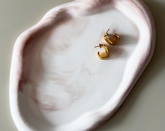 Könnte beinhalten: Ein weiß-rosa marmorierter Dekoteller mit zwei kleinen goldenen Creolen. Der Teller hat eine organische Form und eine glatte Oberfläche. Die Ohrringe sind schlicht und elegant, mit poliertem Finish. Ideal für Schmuck oder kleine Gegenstände.