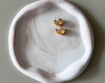 Könnte beinhalten: Ein Paar goldfarbene Creolen liegt auf einem weiß-rosa marmorierten Dekotablett. Das Tablett hat eine organische, unregelmäßige Form. Die Ohrringe sind klein und haben ein geschwungenes Design. Das Tablett besteht aus einem glatten Material.