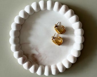 Könnte beinhalten: Ein weißes, rundes Dekotablett mit Perlenrand, das zwei goldfarbene Creolen enthält. Das Tablett hat ein marmorähnliches Muster in der Mitte. Die Ohrringe sind klein und haben eine unregelmäßige Form.