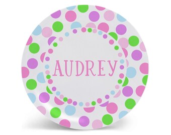 May include: A white round plate with a pink, green, and blue polka dot pattern. The plate has a white circle in the center with the name "AUDREY" written in pink.