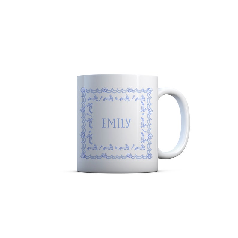 Puede incluir: Taza de cer&aacute;mica blanca con un borde azul con un patr&oacute;n repetido de conejos. El nombre "EMILY" est&aacute; impreso en el centro de la taza.