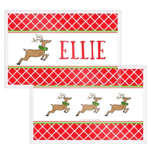 May include: Rectangular Christmas-themed placemats with a red and white diamond pattern border. One placemat features a reindeer illustration and the name "ELLIE" in red. The other placemat has three reindeer illustrations.