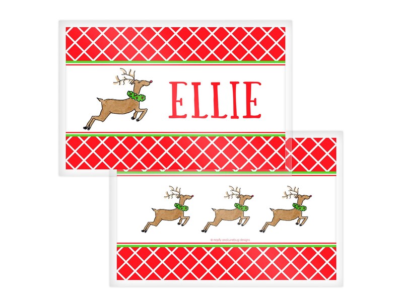 May include: Rectangular Christmas-themed placemats with a red and white diamond pattern border. One placemat features a reindeer illustration and the name "ELLIE" in red. The other placemat has three reindeer illustrations.
