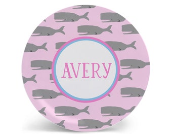 May include: A round, pink plate with a pattern of gray whales. The center of the plate has a white circle with the name "AVERY" in pink letters, surrounded by a blue and pink border. The plate is designed for children.