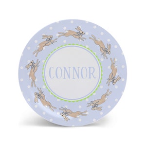 May include: A blue and white personalized plate with a green border and a circle in the center that says "CONNOR". The plate is decorated with a pattern of white dots and brown bunnies with blue bows.