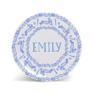 Puede incluir: Un plato de cer&aacute;mica blanco con un borde azul y blanco que presenta un patr&oacute;n repetitivo de conejos, zanahorias y huevos. El centro del plato tiene el nombre "EMILY" escrito en azul.