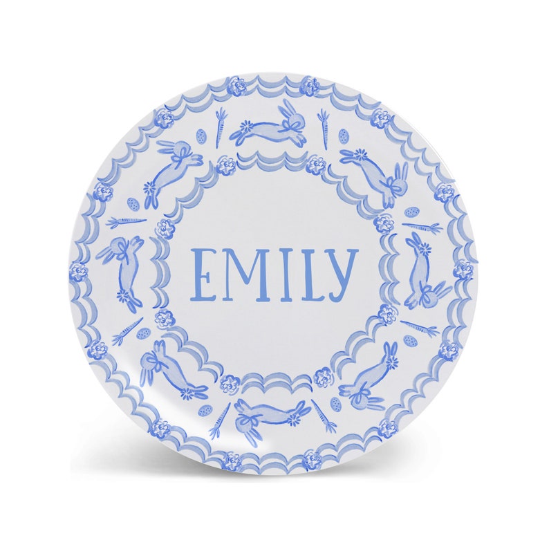 Puede incluir: Un plato de cer&aacute;mica blanco con un borde azul y blanco que presenta un patr&oacute;n repetitivo de conejos, zanahorias y huevos. El centro del plato tiene el nombre "EMILY" escrito en azul.