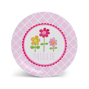 May include: Pink and white children's plate with a pink, yellow, and orange flower design. The plate has a pink and white checkered pattern and the name "Millie" is written in black letters.