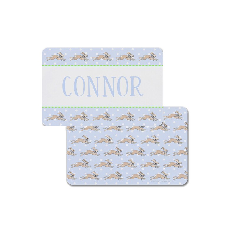 May include: A blue and white placemat with a pattern of brown rabbits running across a blue background with white polka dots. The placemat has a white banner with the name "CONNOR" in green text.