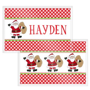 May include: Christmas-themed rectangular placemats with a red and white checkered pattern. Each placemat features a Santa Claus illustration carrying a sack. The name "HAYDEN" is printed in red on one of the placemats.