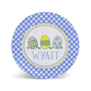 May include: A blue and white gingham patterned plate with a yellow border. The center of the plate features a white circle with three cartoon Easter eggs in a row. The eggs are decorated with yellow, green, and blue colors. The name "WYATT" is written in black letters below the eggs.