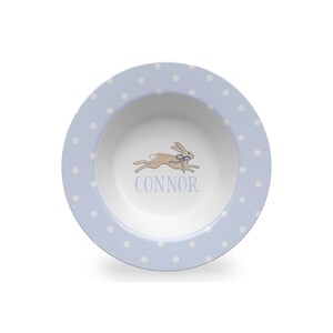 May include: A light blue ceramic bowl with white polka dots. The bowl features a running rabbit illustration and the name "CONNOR" in blue lettering.