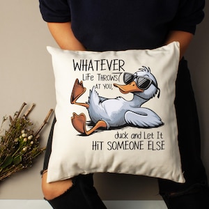 May include: Cream-colored square pillow featuring a cartoon duck wearing sunglasses, reclining with its feet up. The pillow has the text "WHATEVER LIFE THROWS AT YOU, duck and Let It HIT SOMEONE ELSE" in black.