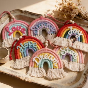 May include: A collection of colorful macrame rainbow wall hangings. Each rainbow features a different word: "Love", "Sunny", "Joy", "Hope", "Dream", and "Stars". The rainbows are made with various colors and have a fringe at the bottom.