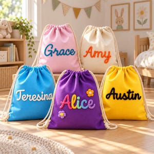 Puede incluir: Cinco mochilas de cordón en rosa, crema, azul, morado y amarillo. Cada bolsa presenta un nombre personalizado en letras de hilo en relieve. La bolsa morada incluye pequeños detalles de flores de ganchillo. Todas tienen cuerdas color crema.