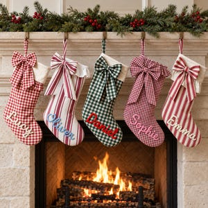 Puede incluir: Cinco calcetines de Navidad colgados de una repisa, cada uno con un lazo de tela y un nombre personalizado. Los calcetines muestran patrones de cuadros vichy rojos y blancos, rayas rojas y blancas, y cuadros vichy verdes y blancos con los nombres Lucy, Oliver, Daniel, Sophie y Emma.