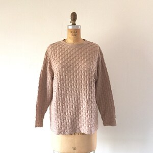 May include: A tan-colored long-sleeved sweater with a textured, geometric pattern. The crew neck sweater is displayed on a vintage dress form with a wire base. The dress form has a wooden top and is set against a plain white background.