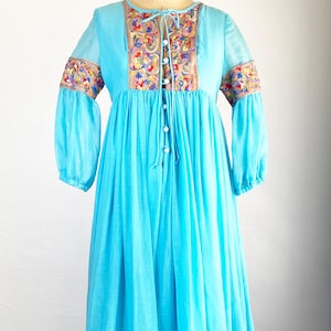 May include: A vintage turquoise maxi dress with long sleeves and a flowing skirt. The dress features colorful embroidered details around the neckline, cuffs, and hem. It has a button-down front and a tie at the neckline, creating a bohemian style.
