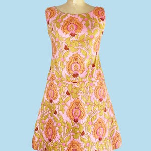 May include: A sleeveless dress featuring a light pink background with a repeating floral and leaf pattern in shades of orange, yellow, and olive green. The garment has a round neckline and a horizontal seam at the waist, creating a structured silhouette.