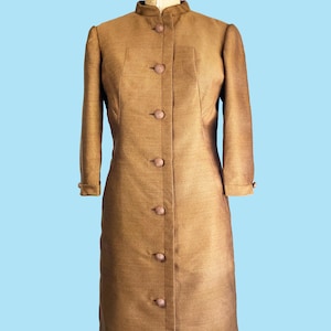 May include: A knee-length, brown coat with a high mandarin collar and long sleeves. The coat features a front button closure with seven matching round buttons and a textured fabric finish. It is displayed on a beige Wolf Model Form mannequin.