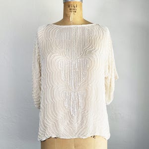 Peut inclure: Haut crème perlé avec un col bateau et des manches courtes. Le chemisier vintage présente un motif symétrique complexe sur le devant. Le vêtement est présenté sur un mannequin vintage avec le texte "MODEL 1965".