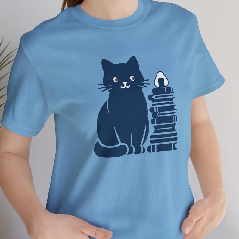 May include: A light blue t-shirt with a black cat sitting next to a stack of books. The cat is smiling and has a white onigiri on top of the stack of books.