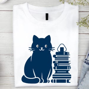 May include: A white t-shirt with a blue silhouette of a cat sitting next to a stack of books. The cat has a happy expression and is looking to the right. The books are stacked on top of each other and have a simple design.