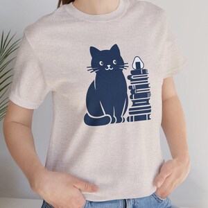 May include: A light gray t-shirt with a black cat and a stack of books printed on the front. The cat is looking to the right and has a stack of books next to it. The books are stacked on top of each other and have a white onigiri on top.