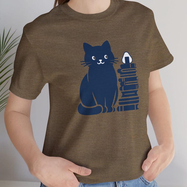 May include: A brown t-shirt with a blue cat and a stack of books. The cat is smiling and has a white onigiri on top of the books.