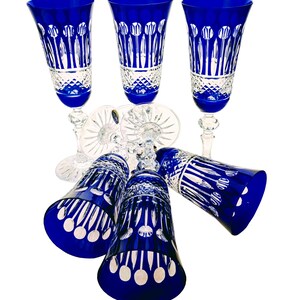 Può includere: Set di sei flûte da champagne in cristallo blu cobalto. Le flûte hanno un gambo e una base trasparenti, con un motivo dettagliato di linee verticali e cerchi. I bicchieri sono disposti su sfondo bianco.