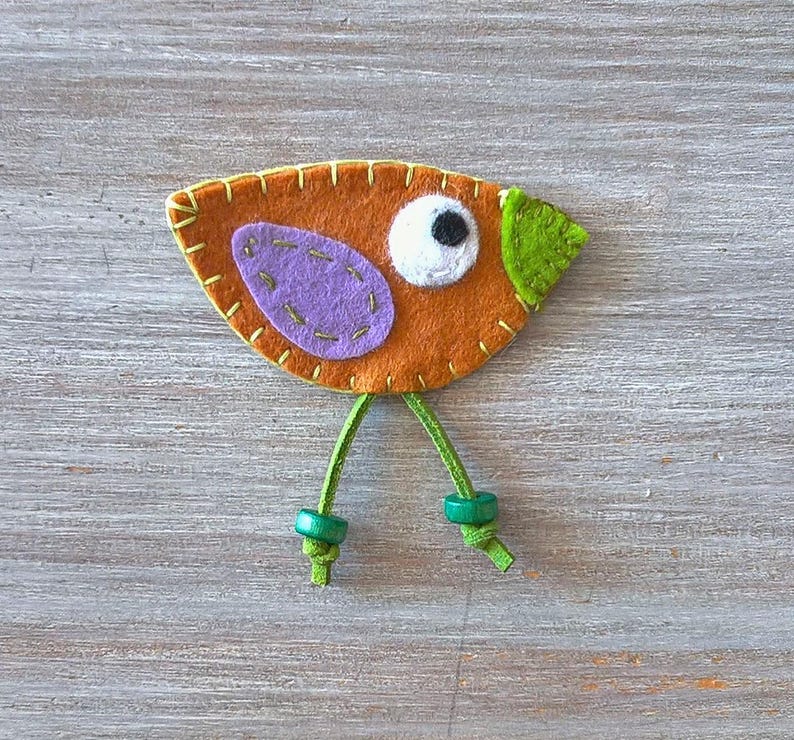 May include: A felt bird ornament with orange, purple, green and white accents. The bird has a white eye with a black pupil and green legs with wooden beads.