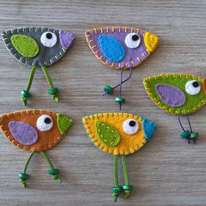 May include: Five felt bird ornaments with different coloured bodies and wings. The birds have green felt legs and black button eyes.