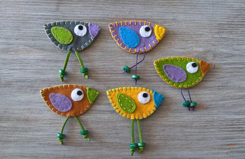 May include: Five felt bird ornaments with different coloured bodies and wings. The birds have green felt legs and black button eyes.