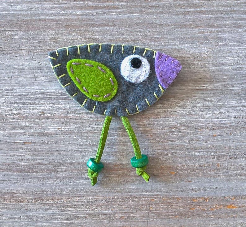May include: A grey felt bird with green felt accents and a purple felt beak. The bird has a white felt eye and green felt legs with green felt buttons at the ends.