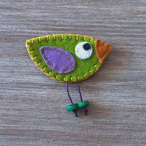 May include: A green felt bird with a purple felt wing, a white felt eye with a black pupil, and an orange felt beak. The bird has green felt legs with purple thread and green felt feet.