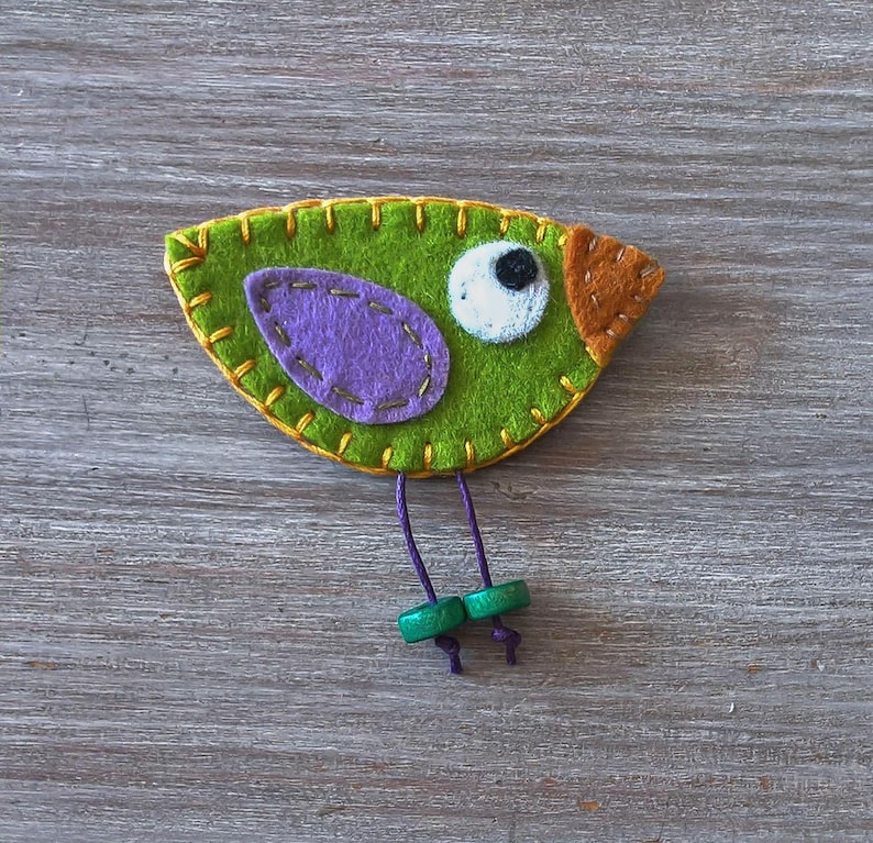 May include: A green felt bird with a purple felt wing, a white felt eye with a black pupil, and an orange felt beak. The bird has green felt legs with purple thread and green felt feet.
