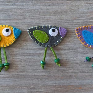 May include: Three felt bird ornaments with green legs and bead accents. The birds are yellow, grey, and purple with green and blue accents.