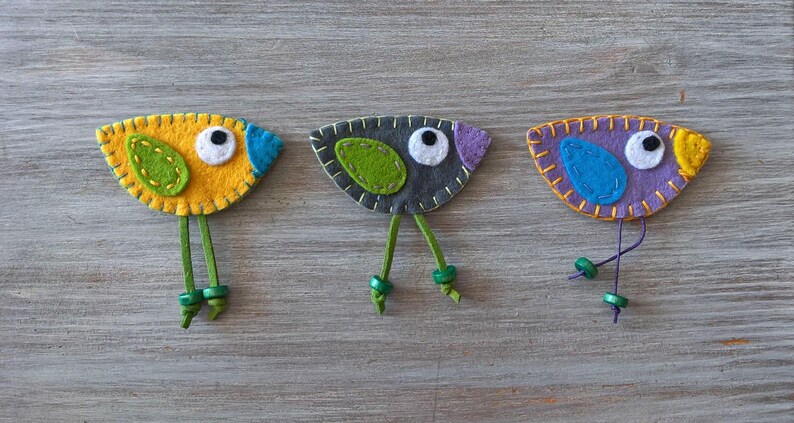 May include: Three felt bird ornaments with green legs and bead accents. The birds are yellow, grey, and purple with green and blue accents.