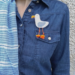Può includere: Una camicia di jeans blu con una sciarpa a righe bianche e blu. Una spilla a forma di gabbiano in feltro bianco con piedi arancioni è appuntata sulla tasca della camicia.