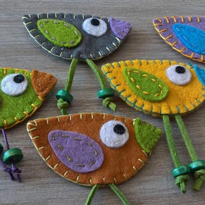 May include: Five felt bird ornaments with green, yellow, orange, purple, and blue accents. Each bird has a white eye and a green stem.