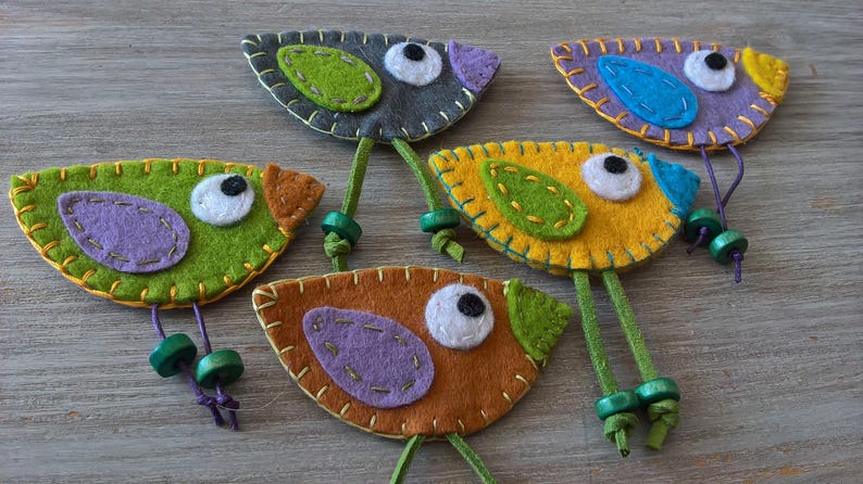 May include: Five felt bird ornaments with green, yellow, orange, purple, and blue accents. Each bird has a white eye and a green stem.