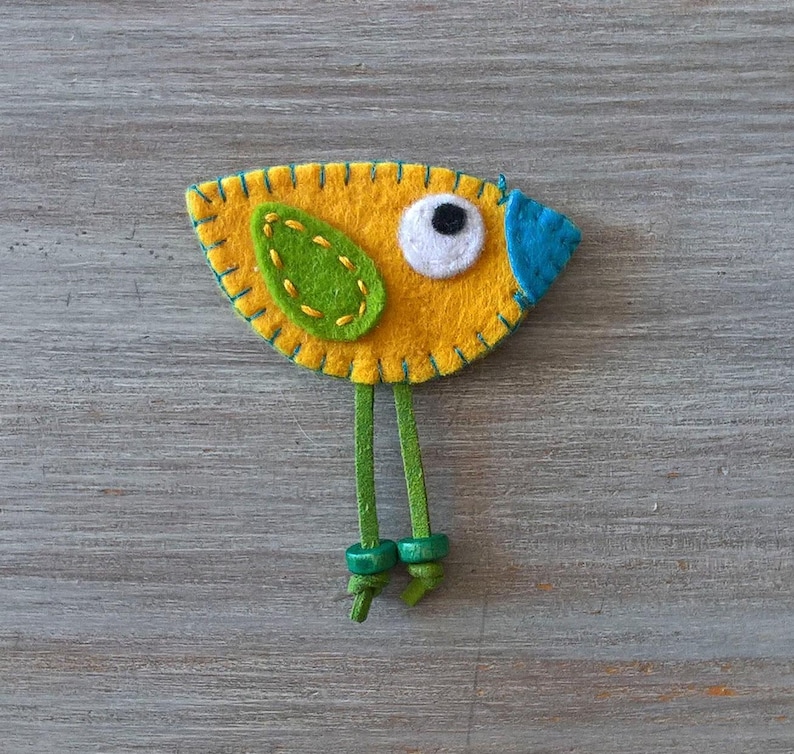 May include: A felt bird ornament with a yellow body, green wing, blue beak, and white eye. The bird has green felt legs with brown buttons.