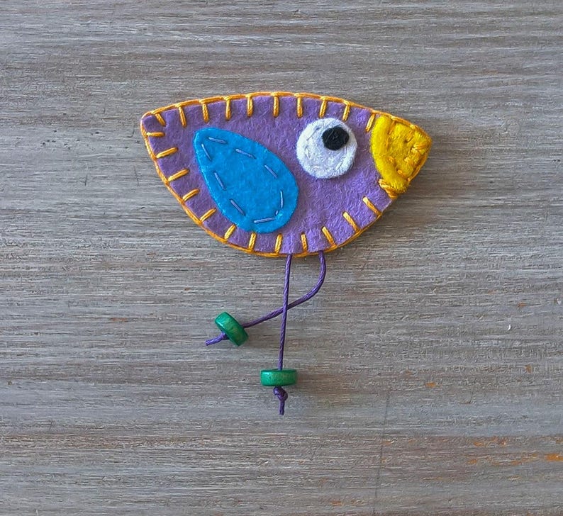 May include: Purple felt bird with a yellow beak, blue wing, and white eye. The bird has green beads on its legs and is attached to a purple string.