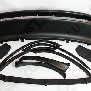 Op de afbeelding: Een verzameling zwarte auto-onderdelen, waaronder een bumper, sideskirts en diverse sierlijsten. De onderdelen zijn geplaatst tegen een witte achtergrond, waarbij hun vorm en ontwerp worden benadrukt. De afbeelding benadrukt de strakke, aerodynamische stijl van de componenten.