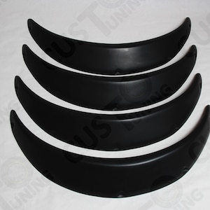 Puede incluir: Cuatro extensiones de paso de rueda de plástico negro para un coche. Las extensiones están apiladas una encima de la otra, con la más grande en la parte inferior y la más pequeña en la parte superior.